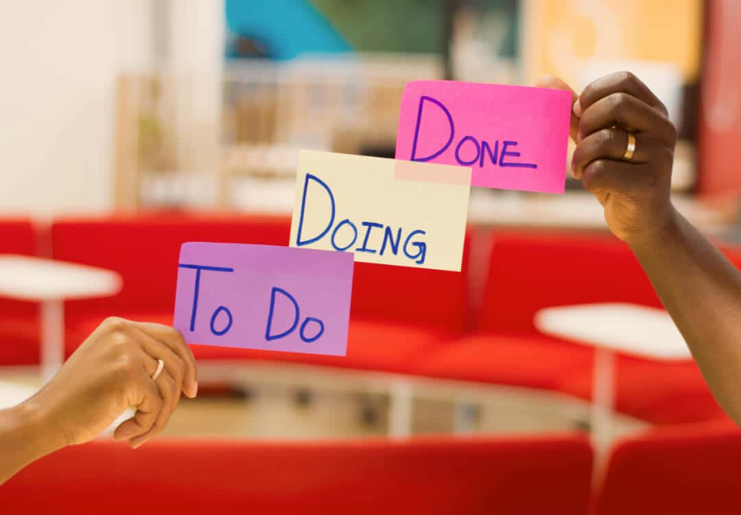 Zwei Hände von unterschiedlichen Personen, die drei Post-It Aufkleber mit der Aufschrift: "To Do, Doing, Done" halten.